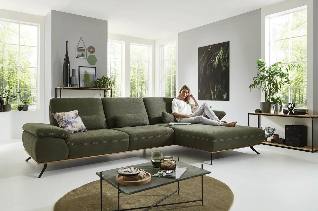 Modernes Wohnzimmer mit grünem Ecksofa, Couchtisch aus Glas, Pflanzen und großen Fenstern. Frau entspannt auf der Couch in stilvoll eingerichteter Wohnlandschaft mit viel Tageslicht